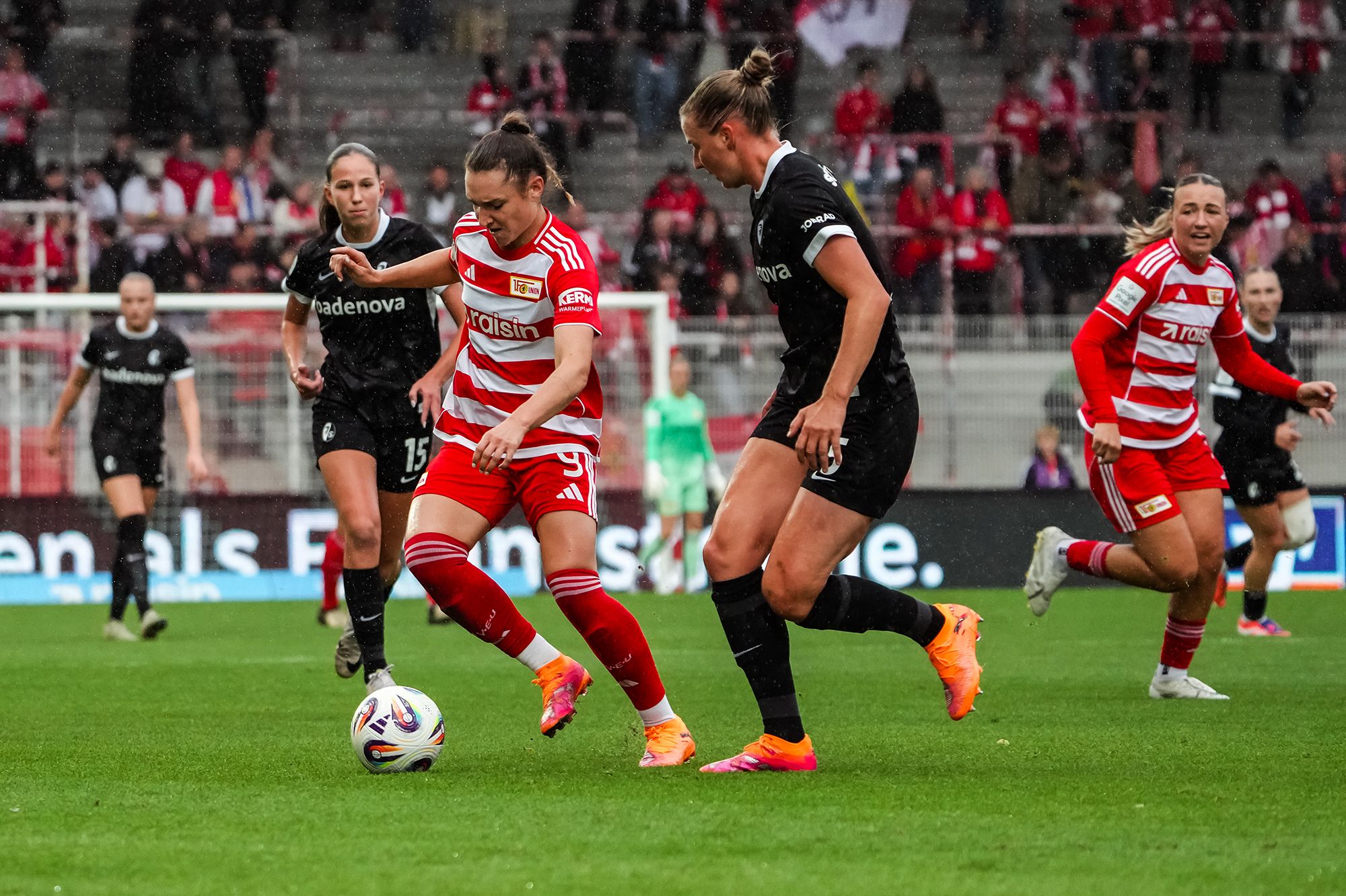 Spielszene eines Frauenfußballspiels mit Spielerinnen in roten und schwarzen Trikots auf dem Platz. Zuschauer im Hintergrund.
