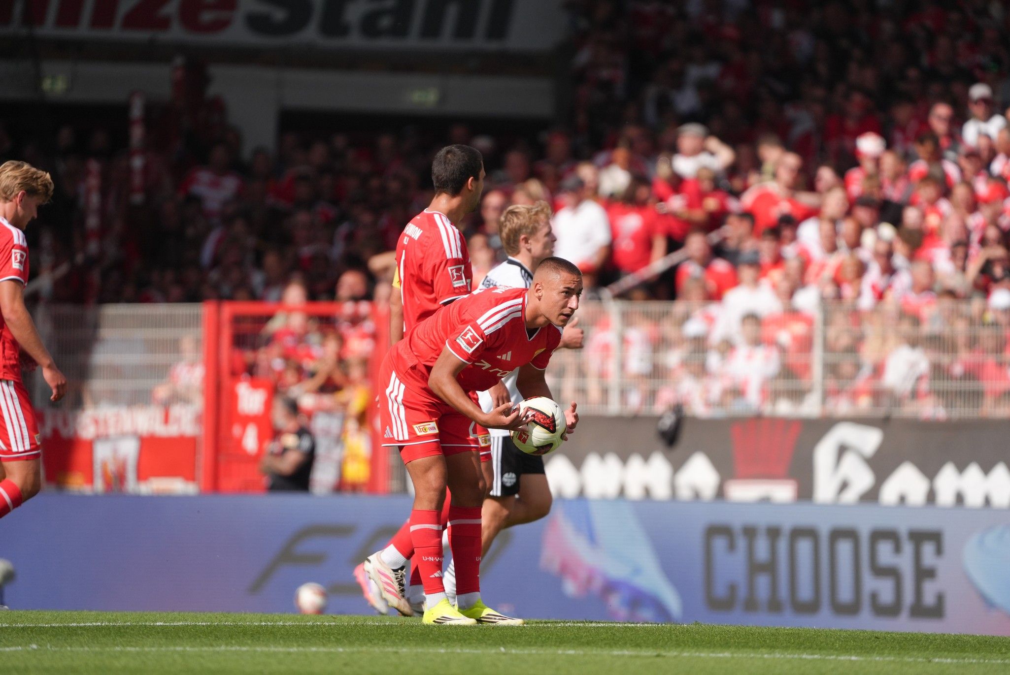 Ein Fußballspieler hält den Ball, um einen Freistoß auszuführen, während andere Spieler in der Nähe stehen. Zuschauer im Hintergrund sind in rot gekleidet.