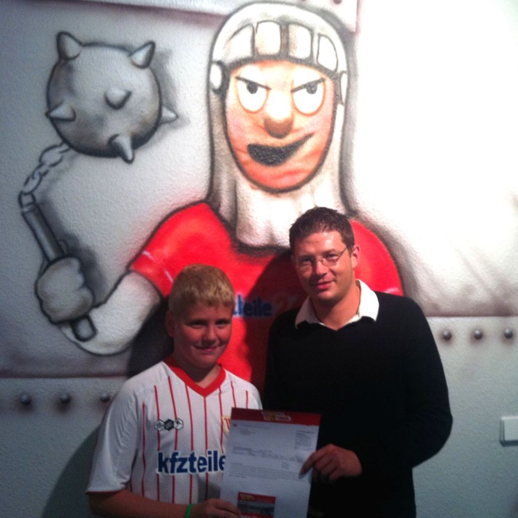 Ein Junge in einem Sporttrikot steht neben einem Mann, der ein Dokument hält; im Hintergrund eine Wandmalerei eines kriegerischen Charakters.