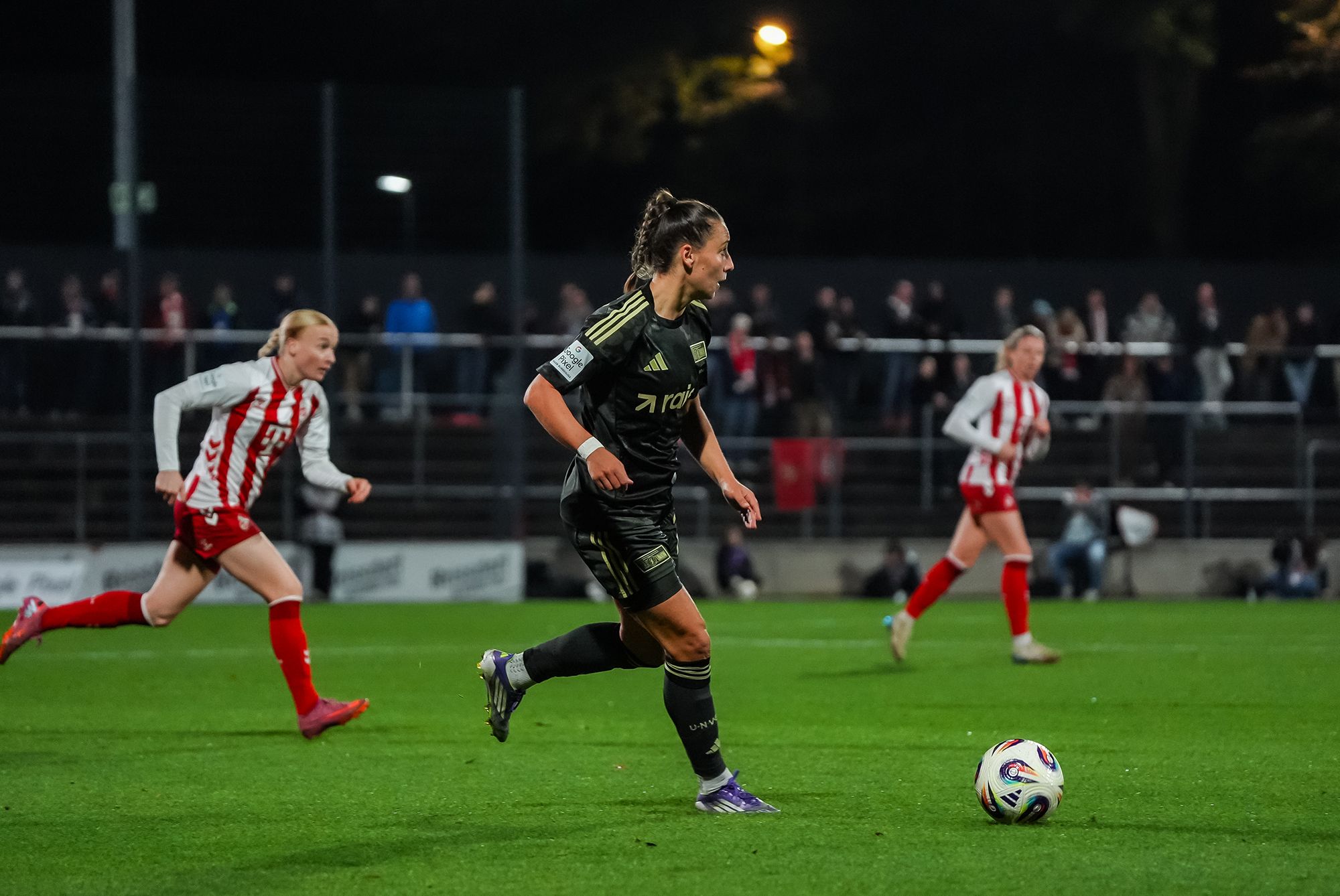 Eine Spielerin in schwarzem Trikot dribbelt den Ball, während eine Gegnerin in rot-weiß im Hintergrund sprintet.