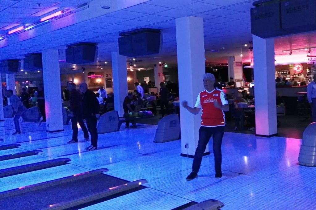 Eine Bowlingbahn mit mehreren Spielern, buntem Licht und einem Mann in einem roten Shirt, der für einen Wurf bereit ist.