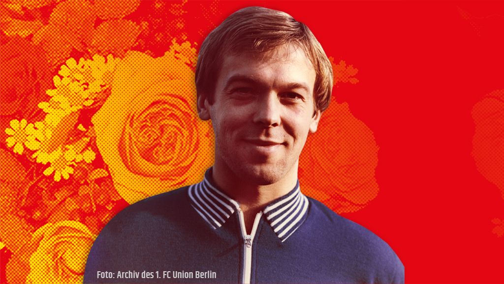 Ein lächelnder Mann mit blonder Frisur trägt ein blaues Oberteil und steht vor einem bunten Blumenmuster.