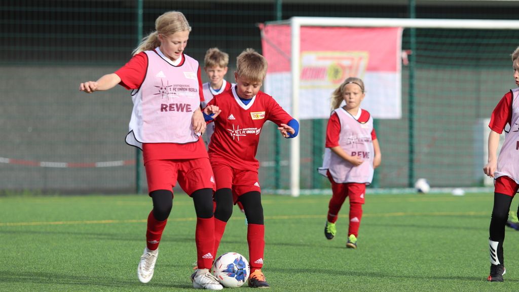 Kinder in roten Fußballtrikots spielen auf einem Kunstrasenplatz und üben das Dribbeln und Passen.