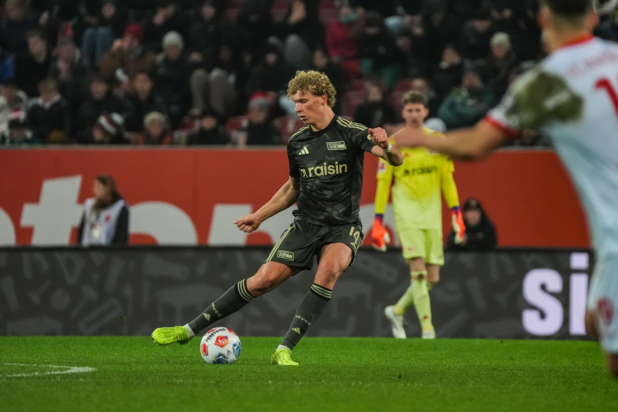 Ein Fußballspieler in schwarzer Trikotfarbe tritt den Ball, während ein Torwart im Hintergrund bereit steht. Zuschauer sind im Stadion sichtbar.