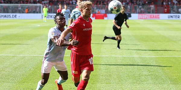 Zwei Spieler spielen Fußball. Einer trägt ein rotes Trikot. Der andere trägt ein graues Trikot. Der Ball fliegt in der Luft.