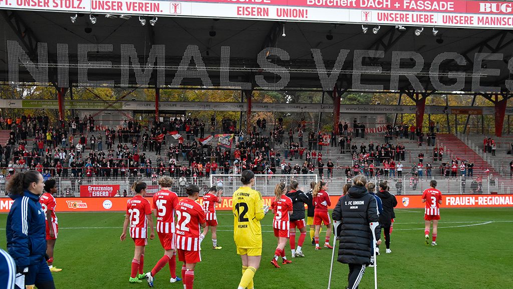Players of a soccer team leave the field, with cheering spectators in the background and large text on the stadium wall.