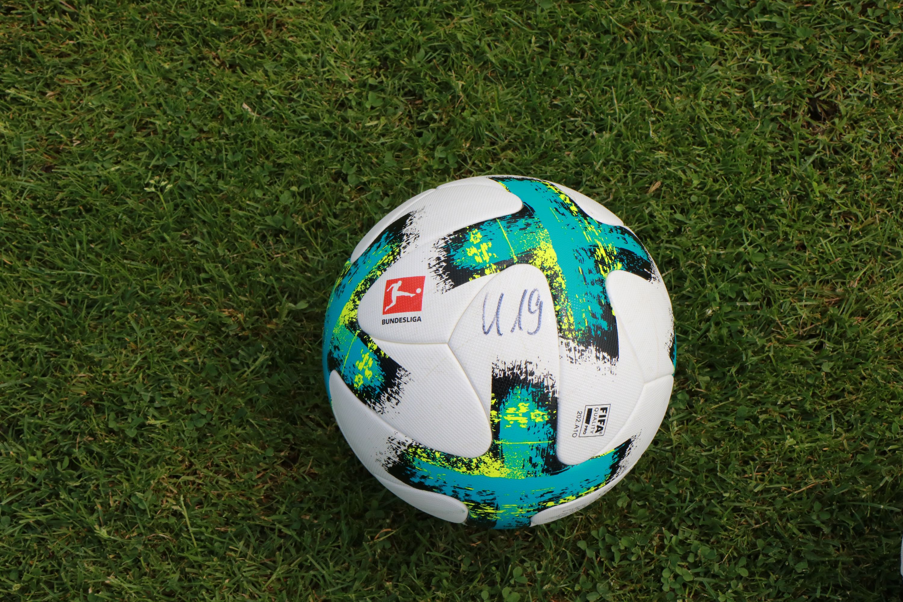 Ein Fußball mit buntem Design und der Aufschrift "U19" liegt auf grünem Gras.