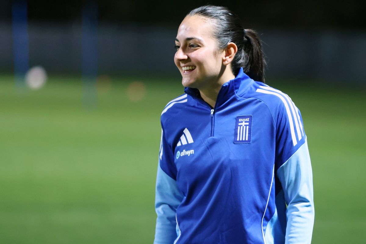 Eine lächelnde Frau in einem blauen Trainingsanzug mit Griechenland-Flagge steht auf einem Fußballplatz.