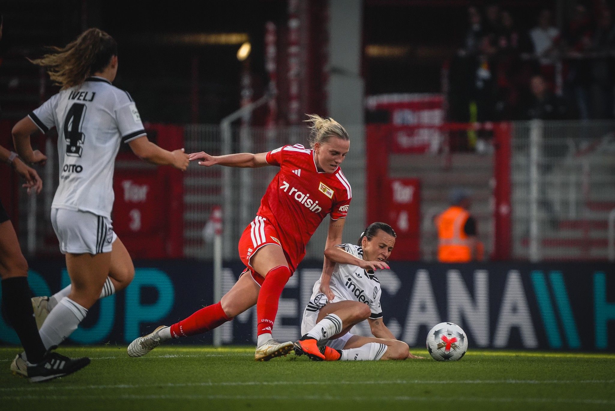 Eine Spielerin in roter Trikotauswahl fällt auf den Boden, während eine gegnerische Spielerin in weiß versucht, den Ball zu erreichen.