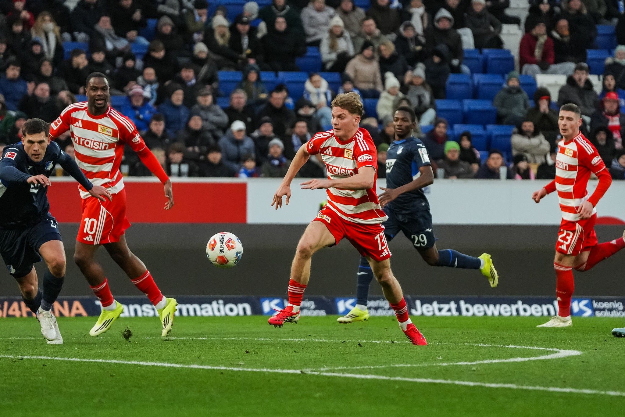 Spielszene mit mehreren Fußballspielern auf dem Platz, einer Spieler in rot-gestreiftem Trikot versucht, den Ball zu spielen.