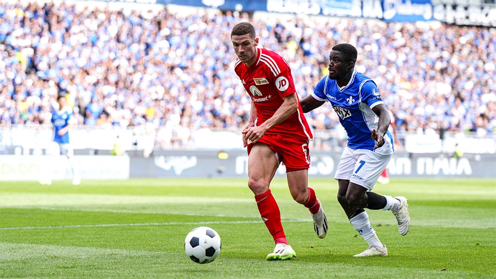A soccer player in a red jersey dribbles the ball while a player in a blue jersey chases him. Fans are visible in the background.