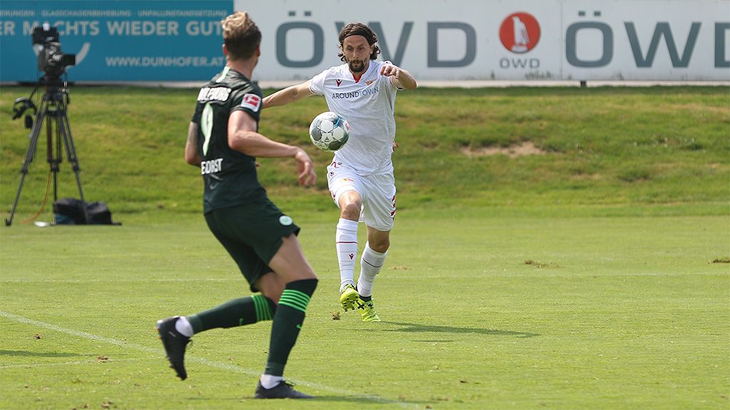 A soccer player in a white jersey is dribbling towards the ball, while an opponent in a green jersey stands opposite him.