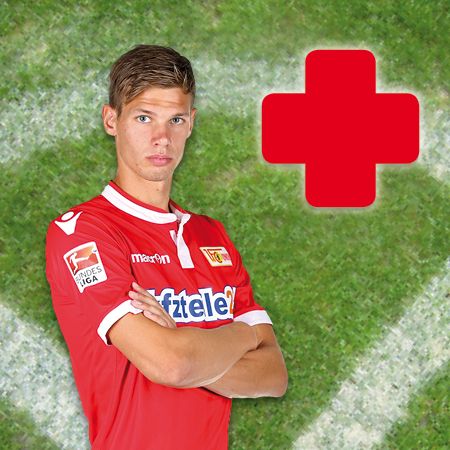 Ein junger Fußballspieler in einem roten Trikot steht mit verschränkten Armen vor einem grünen Hintergrund mit einem roten Pluszeichen.