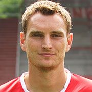 Ein junger Mann mit blondem Haar, der in einem roten Sporttrikot in die Kamera lächelt. Hintergrund unscharf.