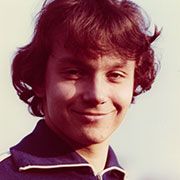 Junger Mann mit lockigem, braunem Haar, der in die Kamera lächelt, vor einem hellen Hintergrund.