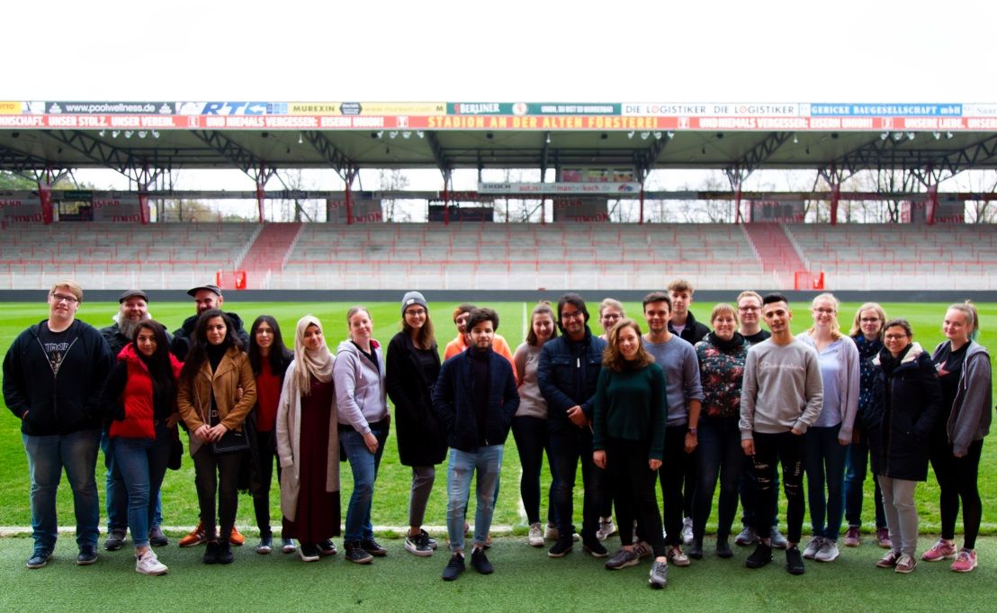 Gruppe von etwa 25 Personen steht lächelnd auf einem Fußballfeld in einem Stadion mit sitzenden Rängen im Hintergrund.