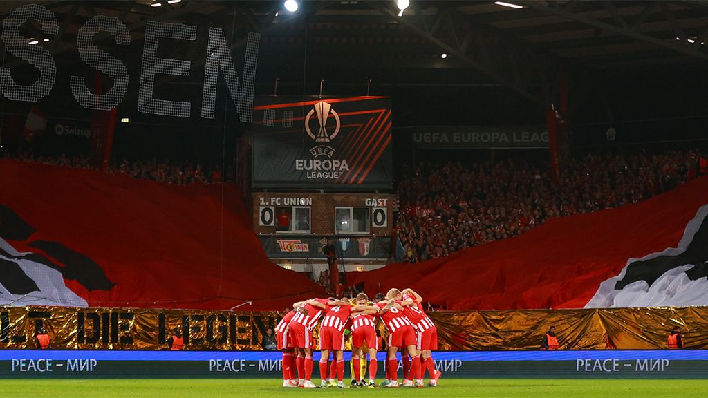 A soccer team stands in a circle preparing for a game, with a large flag in the background and cheering spectators.