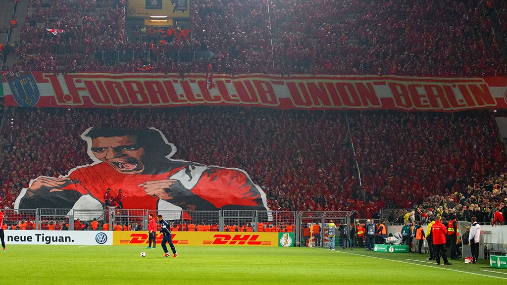 Große Fankurve mit einem Banner des 1. FC Union Berlin und Fans in roten Trikots während eines Spiels.