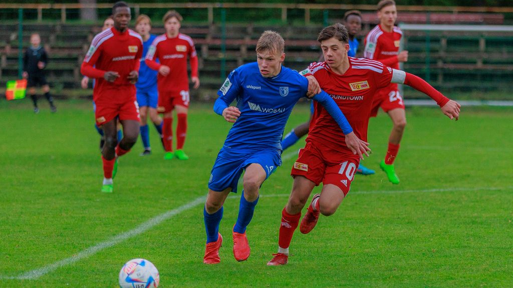 Zwei Fußballspieler in blauen und roten Trikots kämpfen um den Ball auf einem grünen Spielfeld, im Hintergrund weitere Spieler.