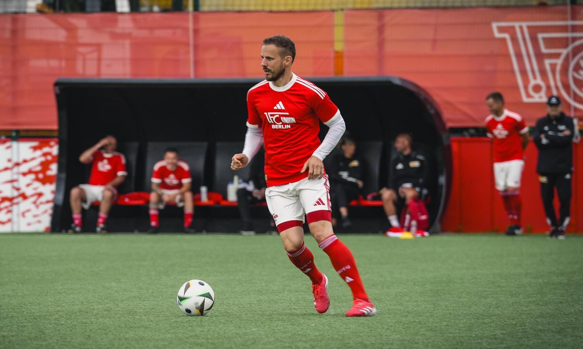 Ein Fußballspieler in rotem Trikot dribbelt den Ball auf einem Kunstrasenplatz. Im Hintergrund sitzen Teamkollegen.