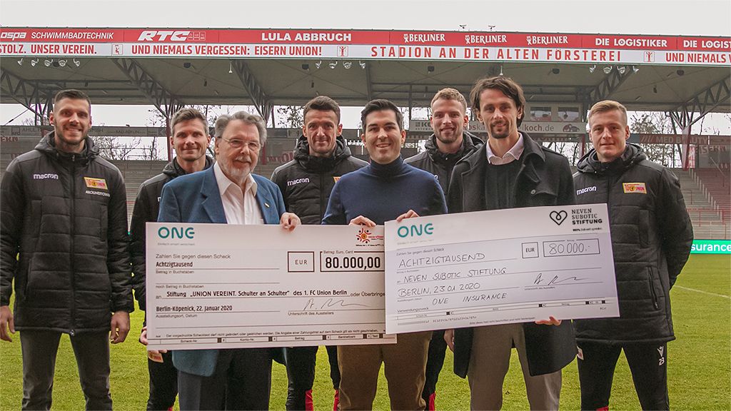 Group scene on a football field, several people present checks for a donation of 800,000 euros.