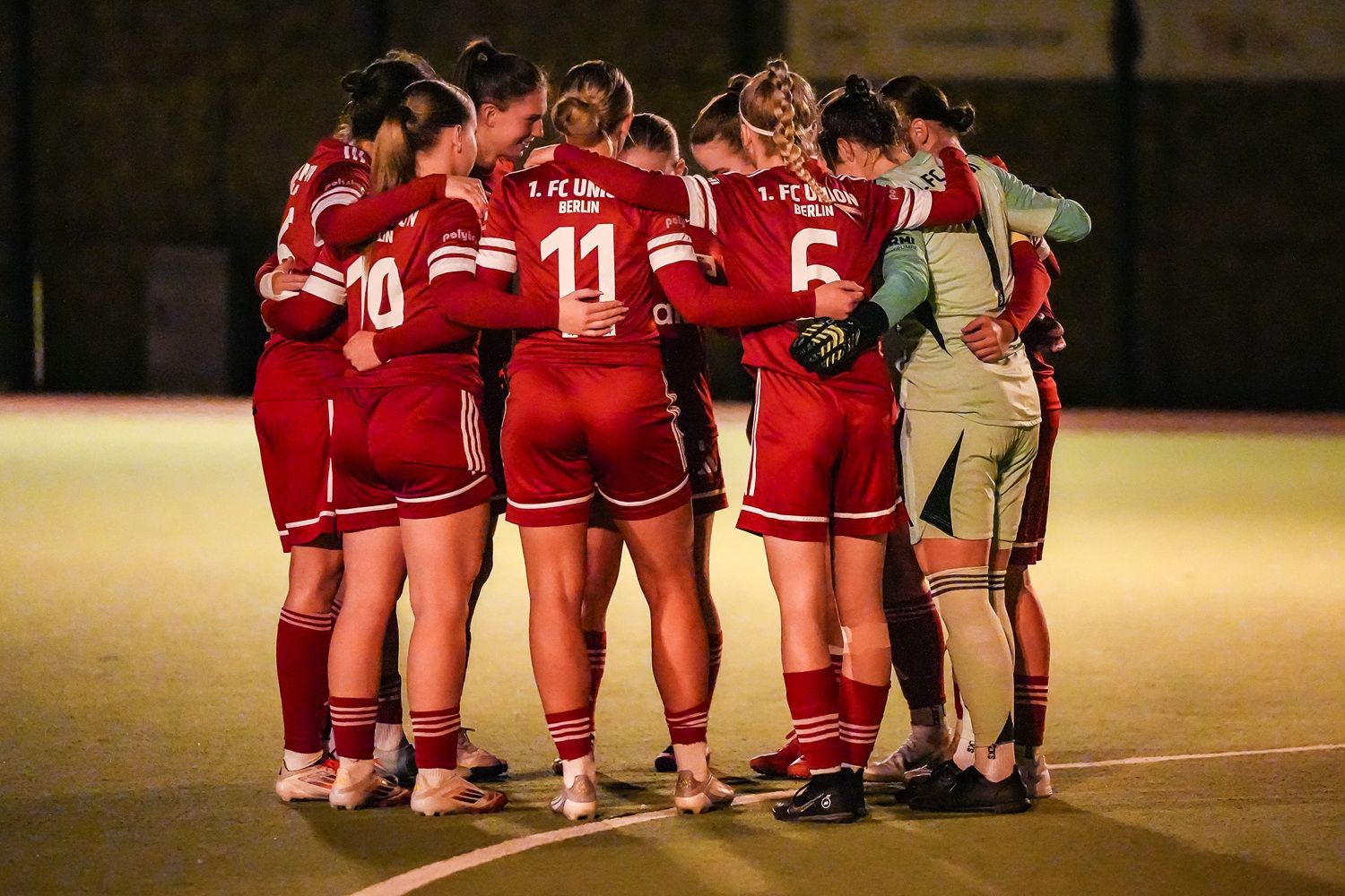 Frauenfußballmannschaft in roten Trikots umarmt sich im Kreis auf dem Spielfeld.