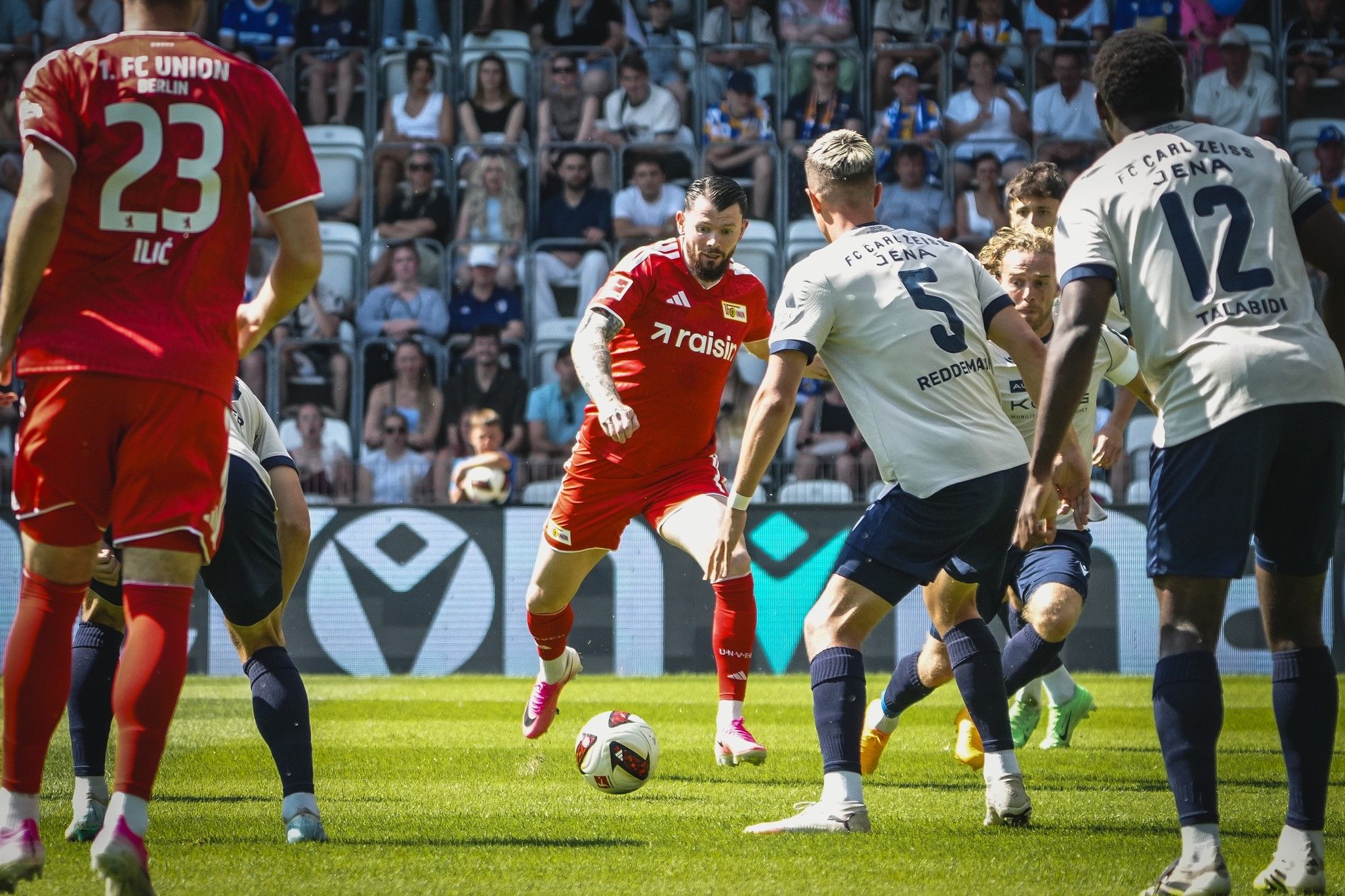 Spielszene während eines Fußballspiels, mit Spielern in roten und blauen Trikots auf dem Feld und Zuschauern im Hintergrund.