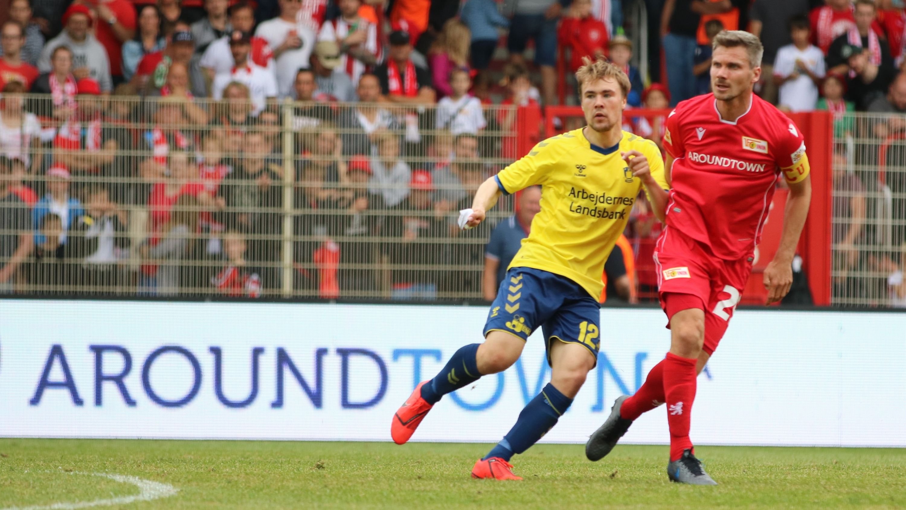 Ein Fußballspieler in gelber Trikotnummer 12 sprintet, während ein Spieler in rotem Trikot mit der Nummer 2 ihn verfolgt. Zuschauer im Hintergrund.