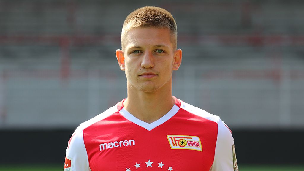 Ein junger Fußballspieler in einem roten und weißen Trikot steht vor einer leeren Stadiontribüne.