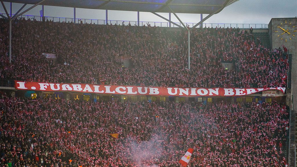Große Menschenmenge in einem Stadion, die ein Banner mit "I. FUSSBALLCLUB UNION BERLIN" hält, umgeben von roten und weißen Farben.