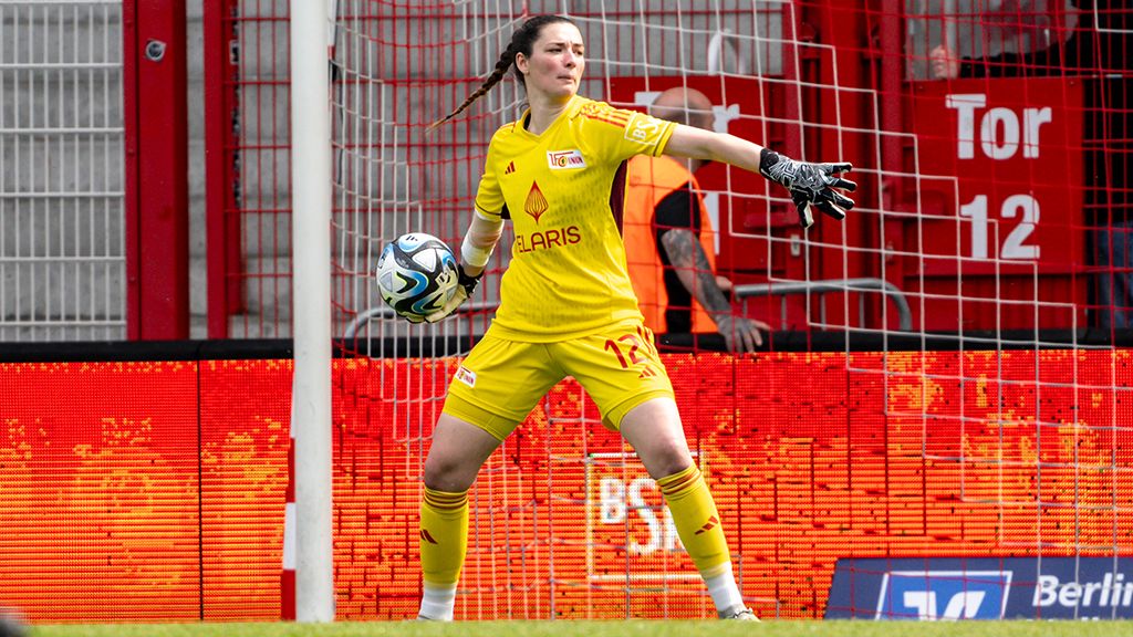 Torhüterin in gelbem Trikot mit Handschuhen, bereitet sich auf einen Spielzug vor, im Stadion mit Zuschauerbereich im Hintergrund.