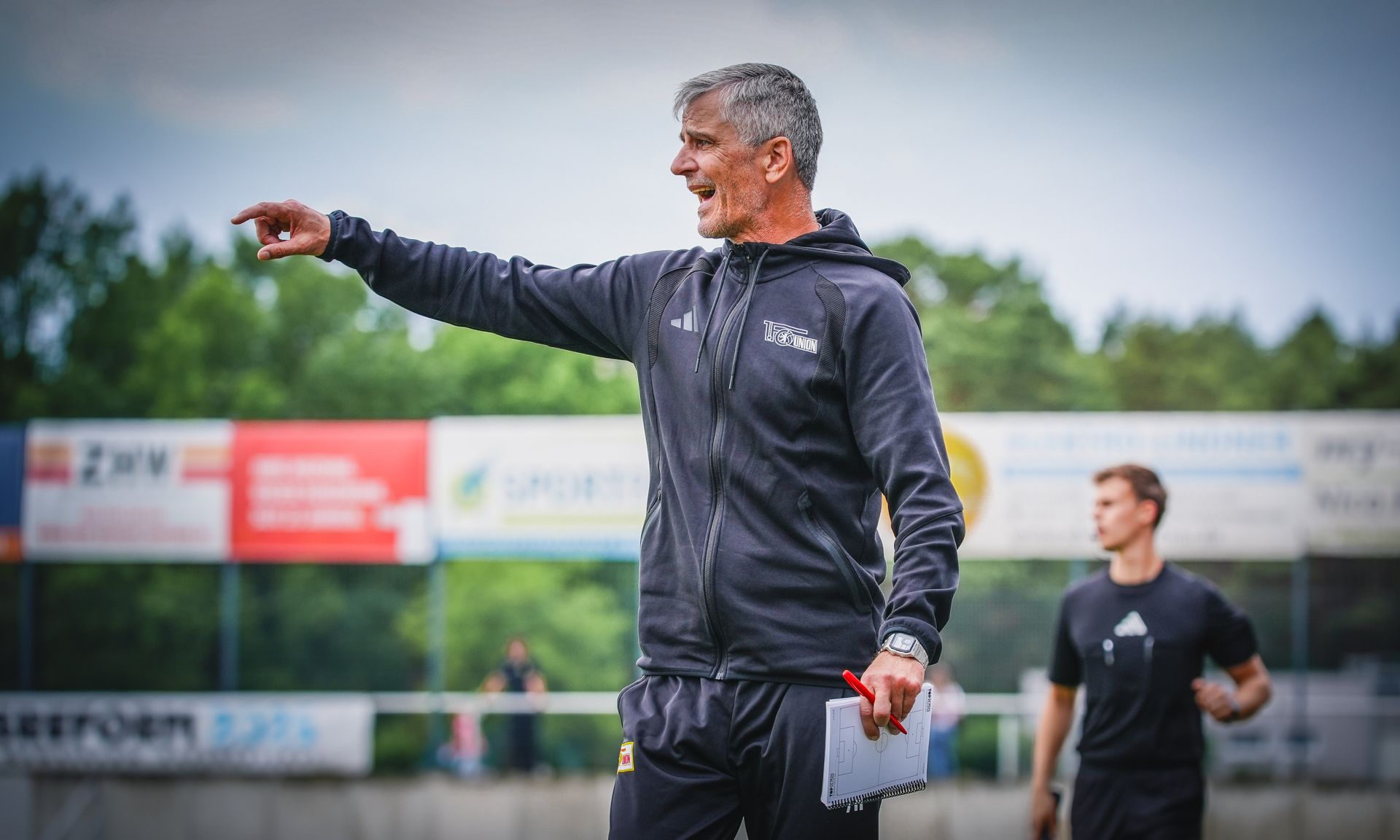 Ein Trainer zeigt mit der Hand auf das Spielfeld und gibt Anweisungen, während ein Spieler im Hintergrund steht.