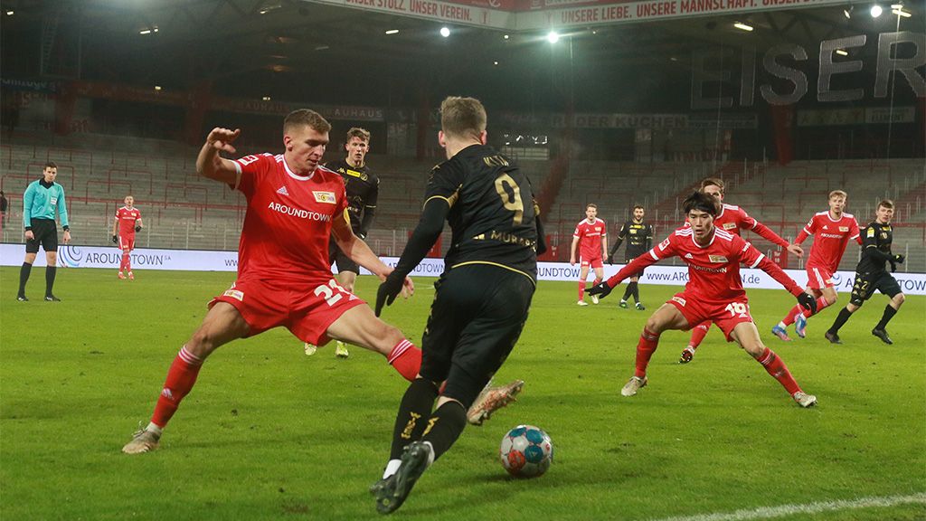 Fußballspiel mit Spielern in roten Trikots, die einen Gegner in schwarzen Trikots attackieren. Im Hintergrund Zuschauer auf der Tribüne.
