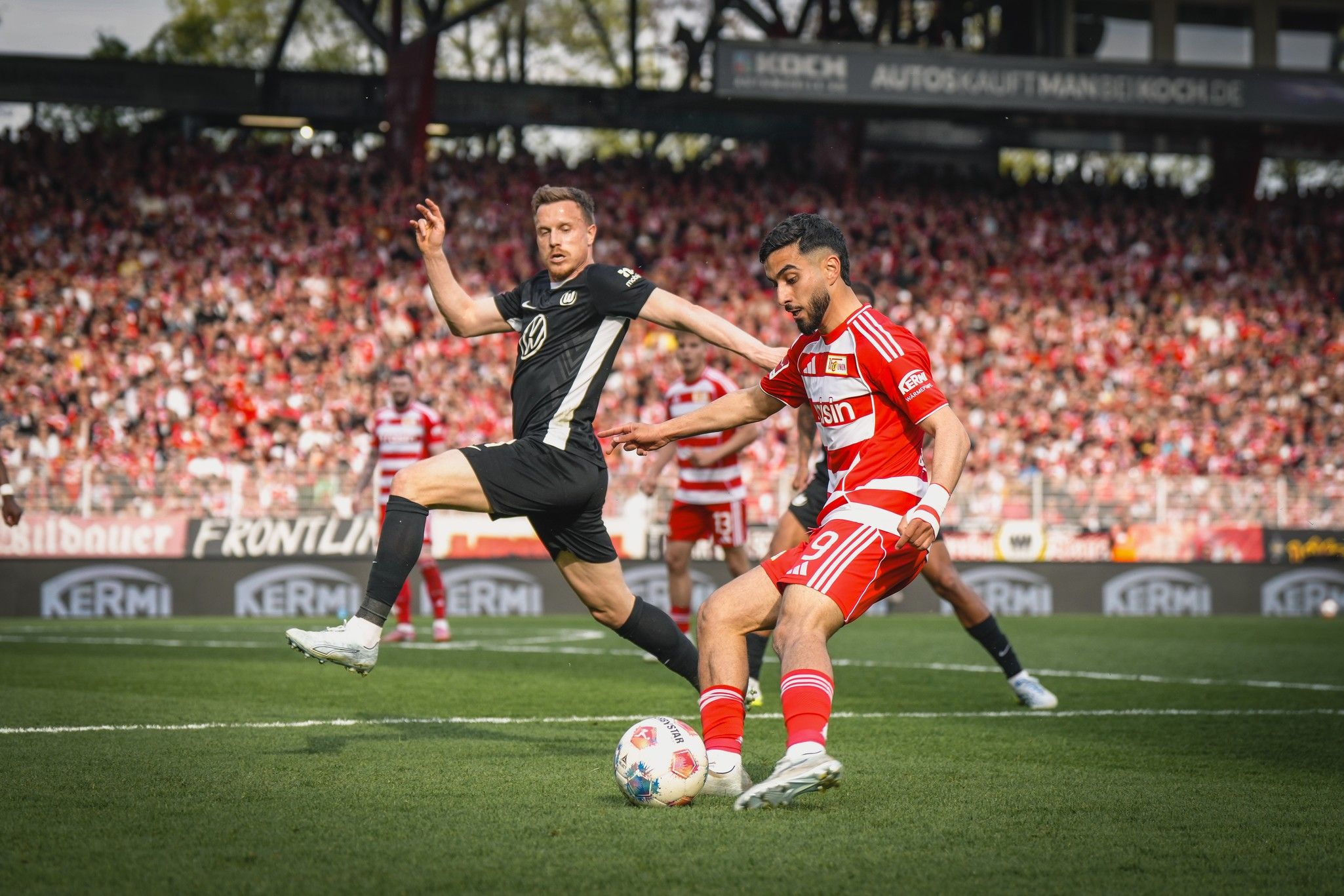 Ein Fußballspieler in rotem Trikot dribbelt den Ball, während ein Gegner in schwarzem Trikot versucht, ihn zu stoppen.