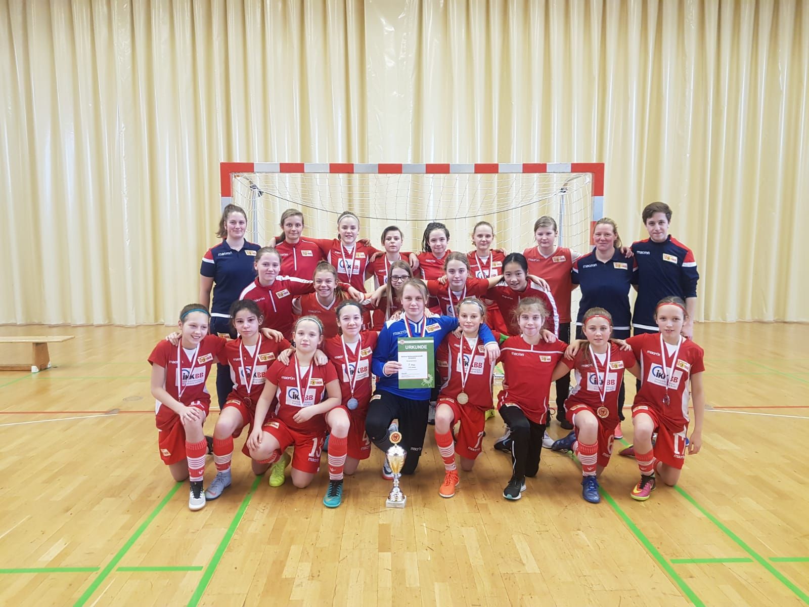 Mädchen-Fußballmannschaft in roten Trikots posiert in einer Halle, einige halten Medaillen und einen Pokal, andere stehen daneben.