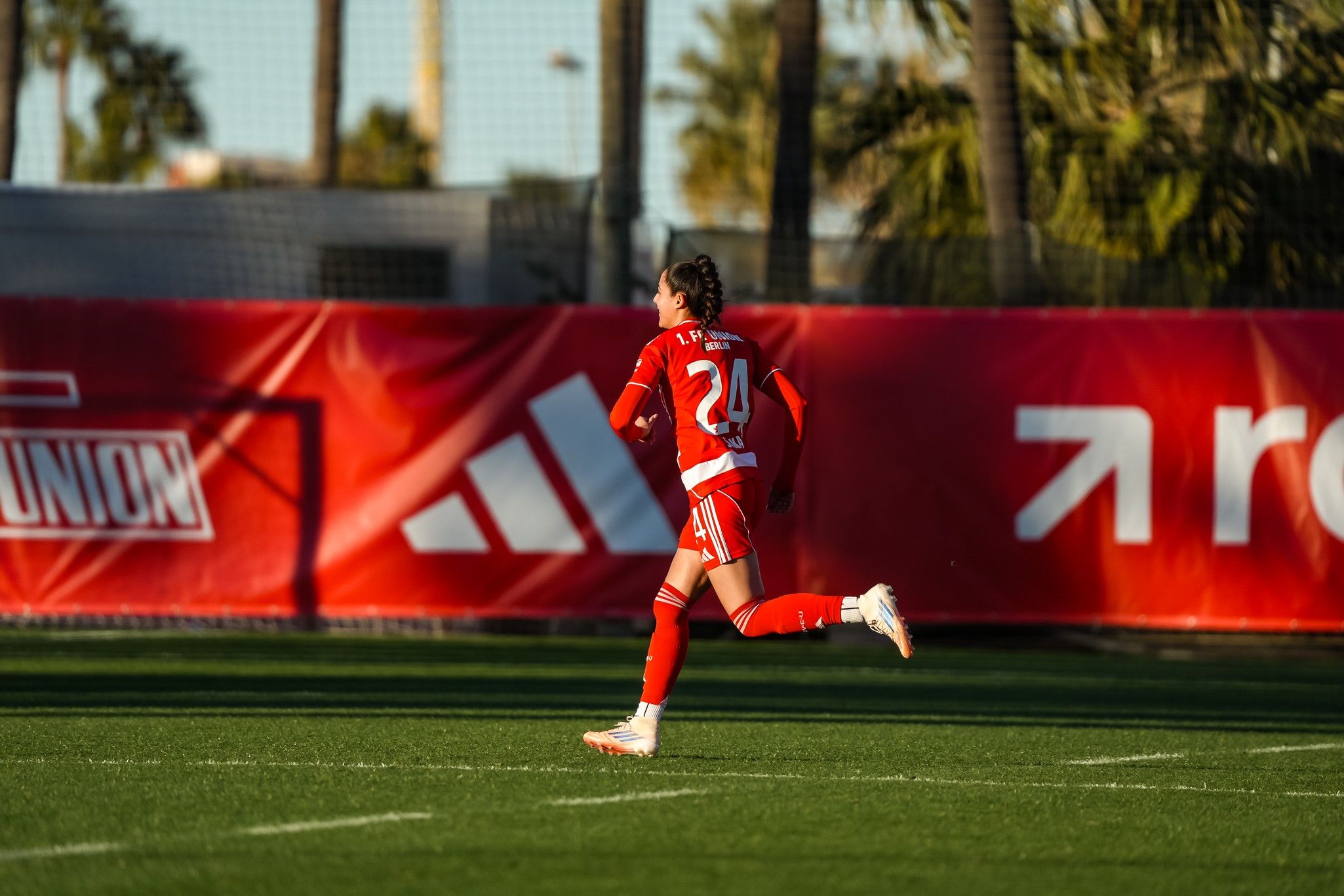 Eine Spielerin in rotem Fußballtrikot rennt auf einem grünen Sportplatz, im Hintergrund sind Palmen und ein Werbebanner zu sehen.