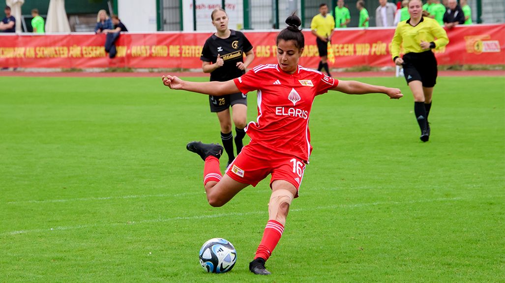Eine Spielerin in rotem Trikot tritt den Ball auf einem Fußballfeld, während andere Spielerinnen im Hintergrund stehen.