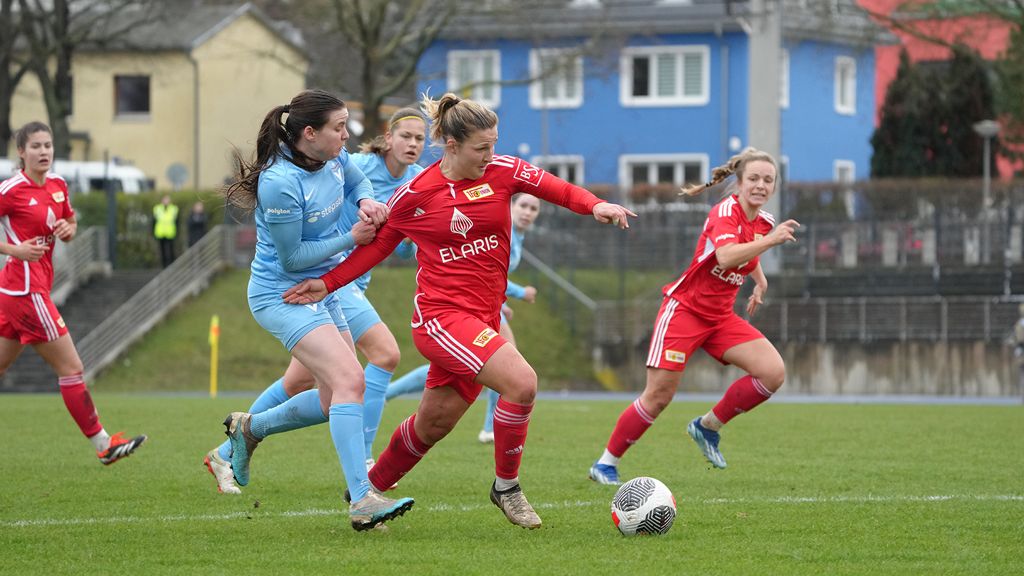 Zwei Frauenfußballerinnen im Wettkampf um den Ball, während eine Spielerin in Blau von einer in Rot bedrängt wird.