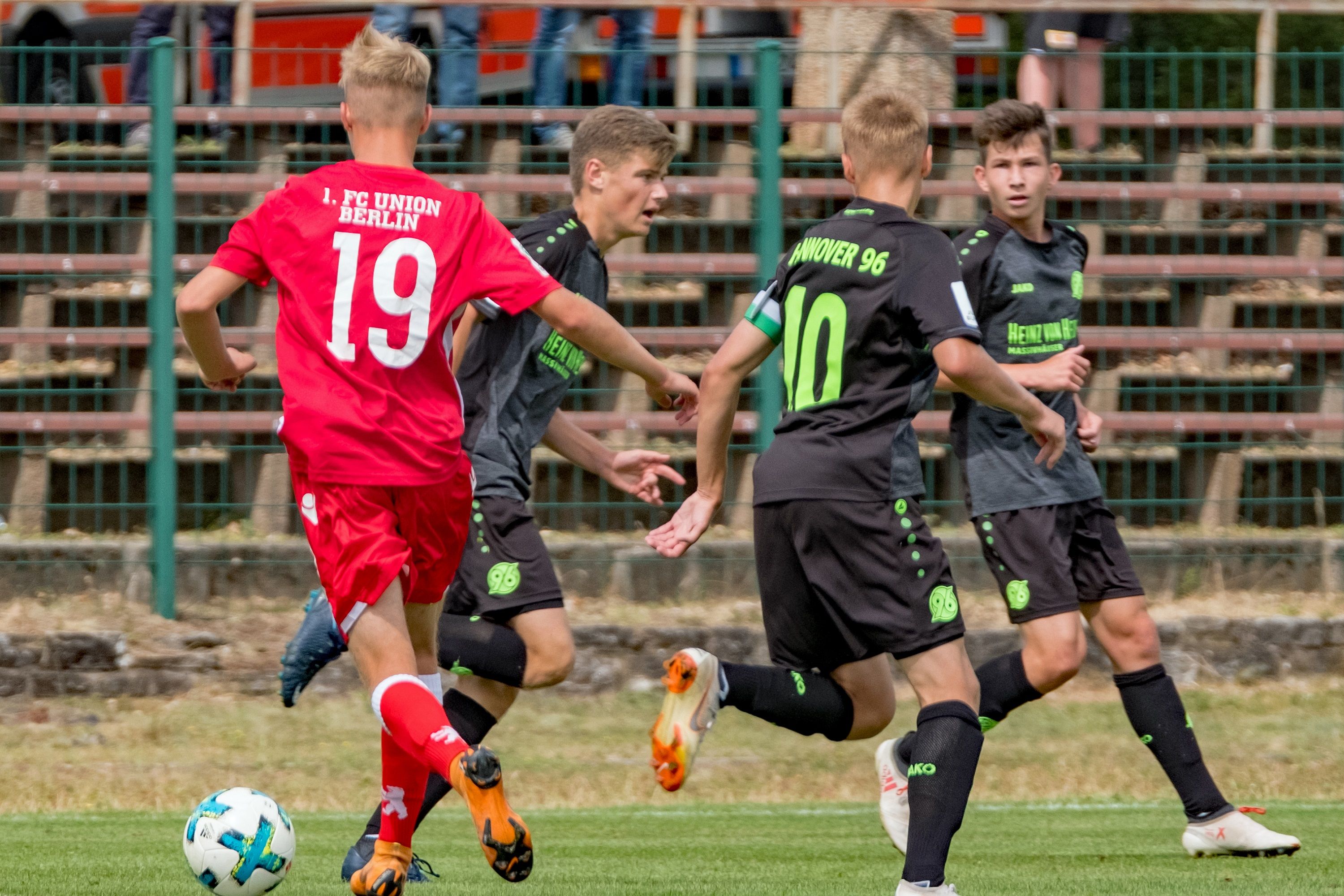 Zwei Fußballspieler tragen schwarz-grüne Trikots. Sie dribbeln. Ein Spieler in rot mit der Nummer 19 spielt gegen sie.