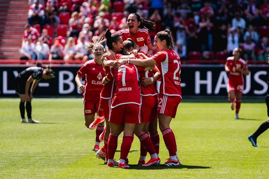 Spielerinnen in roten Trikots feiern einen Torerfolg in einem Stadion voller Zuschauer.