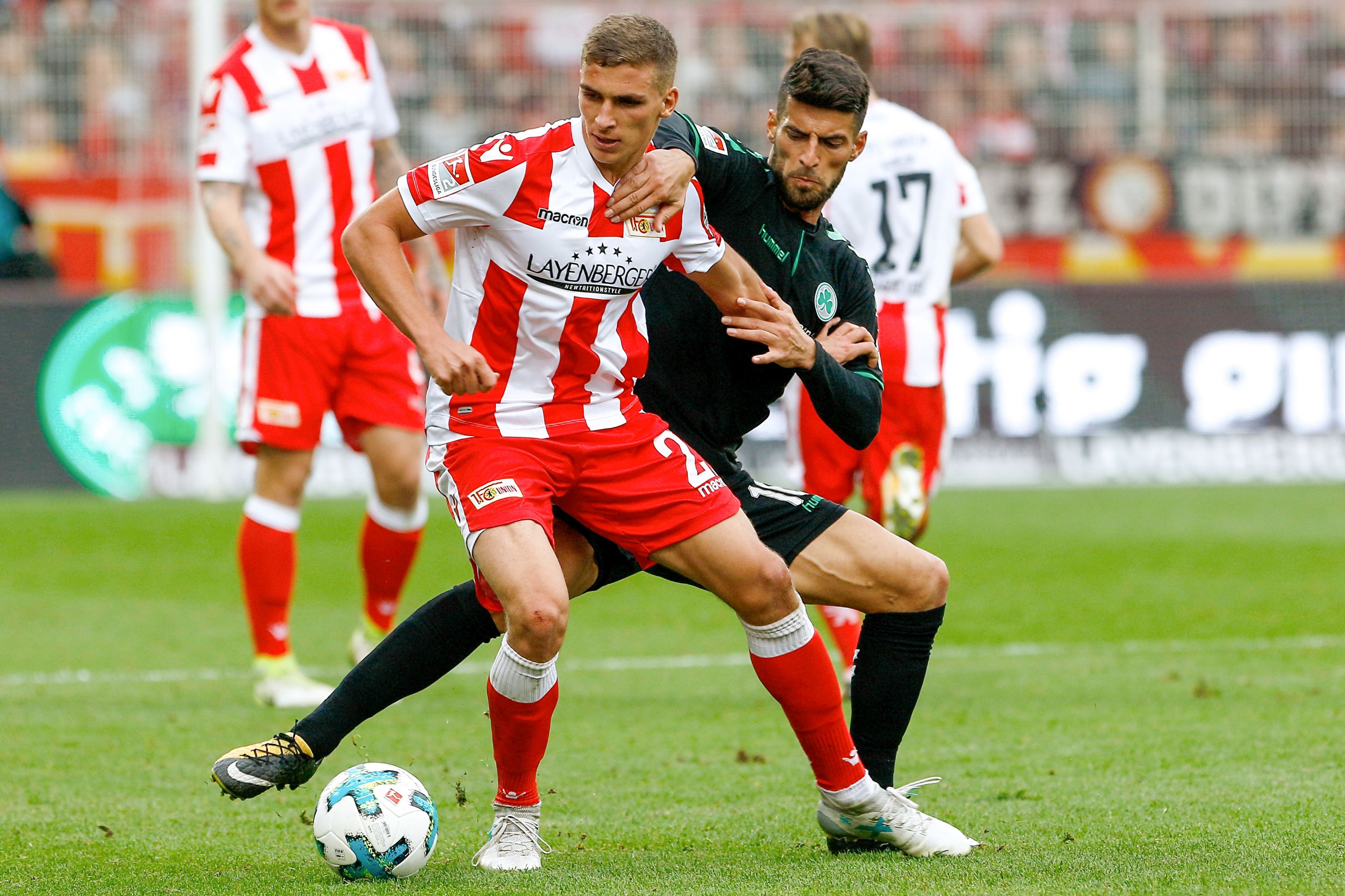 Ein Spieler in rot-weißer Kleidung wird von einem gegnerischen Torwart in einem Fußballspiel von hinten angegriffen.