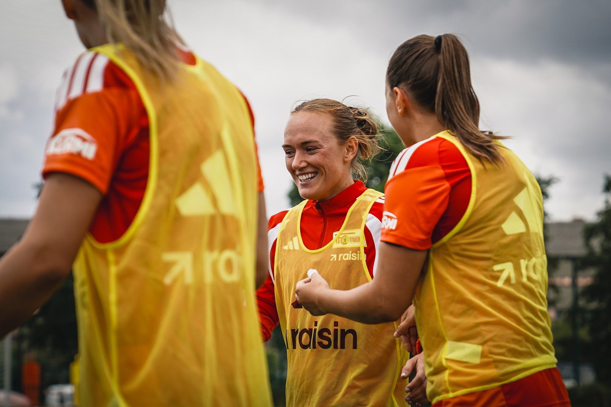 Zwei Frauen in Trainingsanzügen und gelben Überwurfwesten lächeln und interagieren während eines Trainings auf dem Fußballplatz.