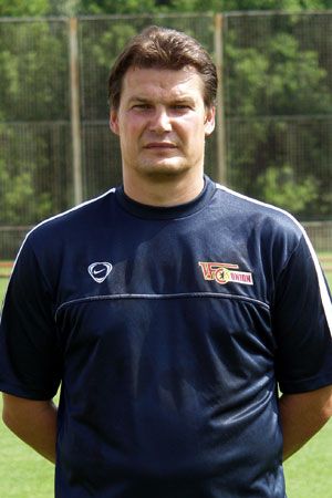 Mann im blauen Sport-Outfit steht auf einem Trainingsfeld mit einem neutralen Gesichtsausdruck.