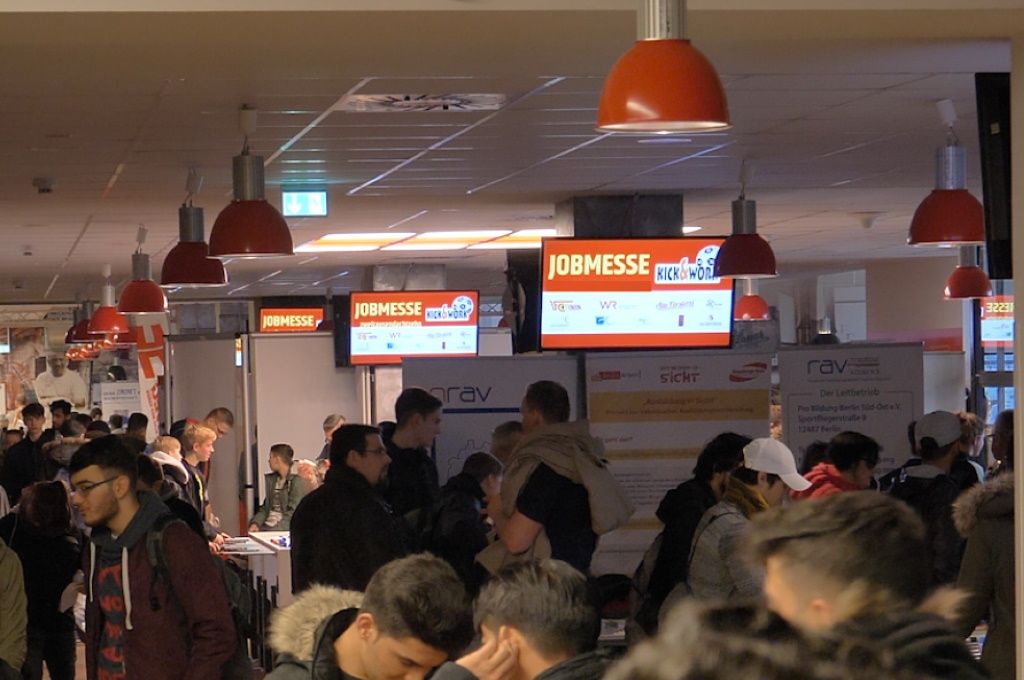Crowd at a job fair, with screens in the background displaying information and advertisements.