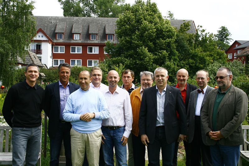 Gruppe von Männern steht im Freien vor einem Hotel, umgeben von Bäumen und Blumen. Sie lächeln und schauen in die Kamera.