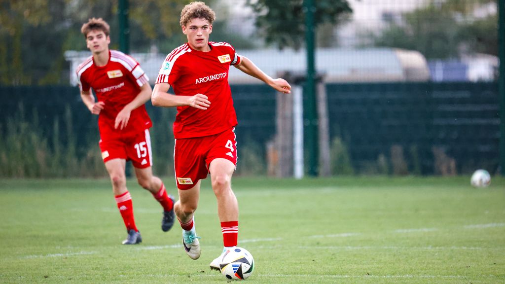 Zwei Fußballspieler in roten Trikots laufen auf einem Rasenplatz, der hintere Spieler verfolgt den Ball.