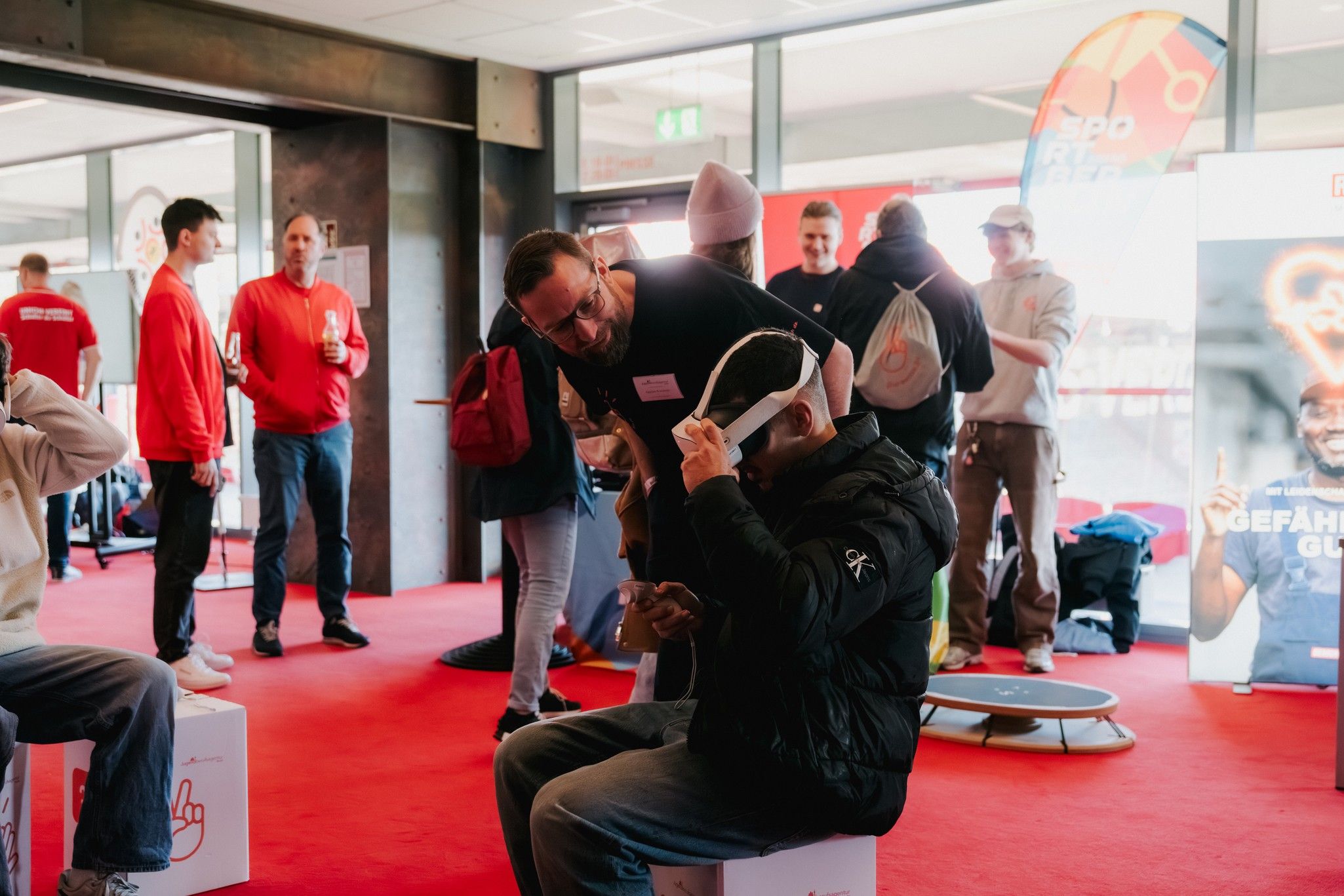 Ein Mann zeigt einem anderen, der einen VR-Helm trägt, etwas. Im Hintergrund stehen weitere Personen in einer farbenfrohen Umgebung.