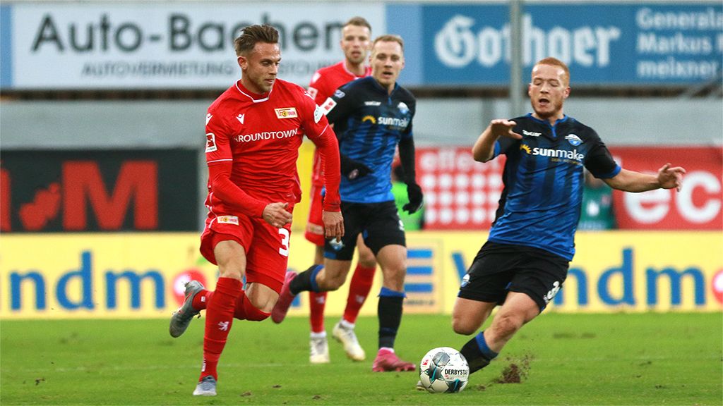Game scene in football with a player in a red jersey dribbling a ball while an opponent in a blue jersey attacks him.
