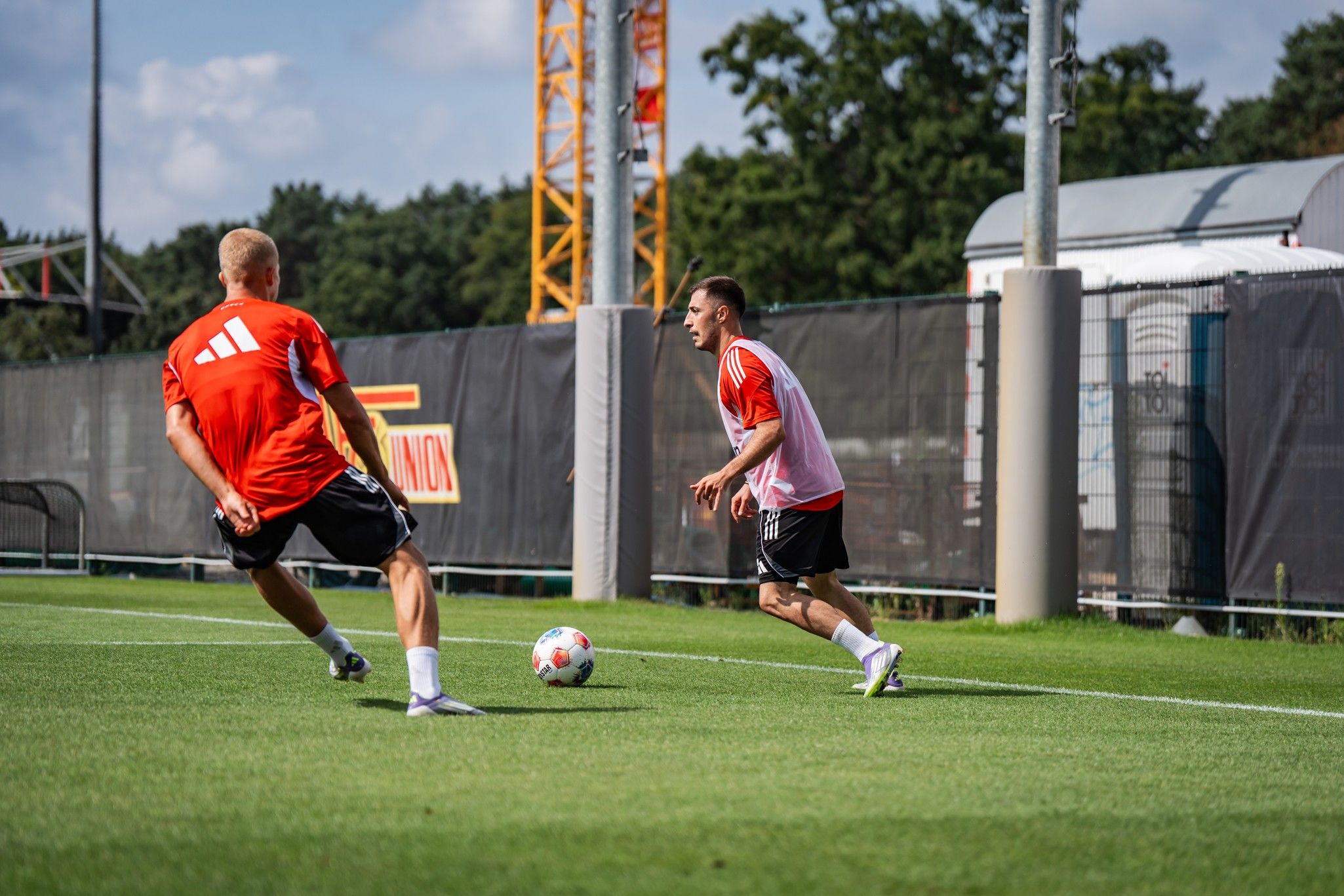 Zwei Fußballspieler in roten Trikots trainieren auf einem Rasenplatz, während sie einen Ball passen.