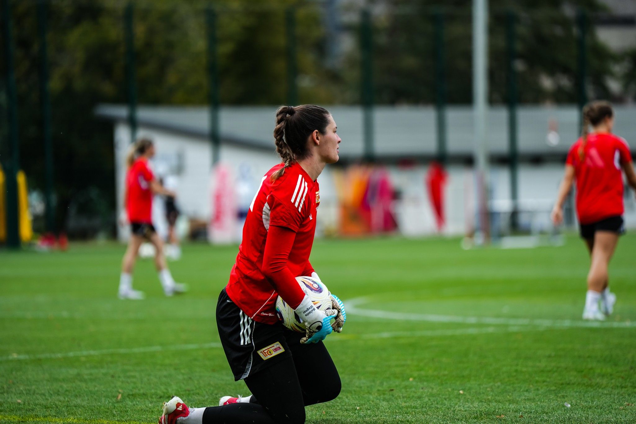 Torfrau im roten Trikot kniet auf dem Fußballplatz und hält den Ball, während im Hintergrund andere Spielerinnen trainieren.